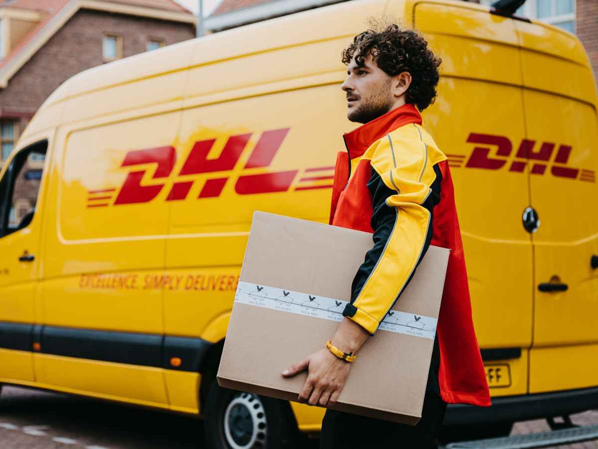 DHL courier carrying a parcel beside a yellow delivery van after new US customs rule freezes shipments over $800