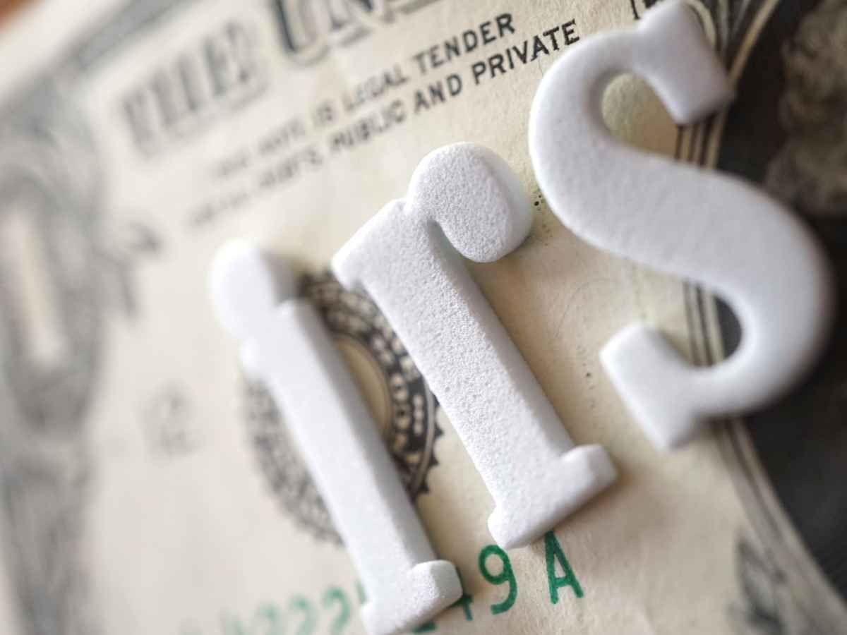 Close-up of white “IRS” letters resting on a U.S. dollar bill, symbolizing potential wage garnishments, bank account freezes, and home foreclosures for unpaid taxes