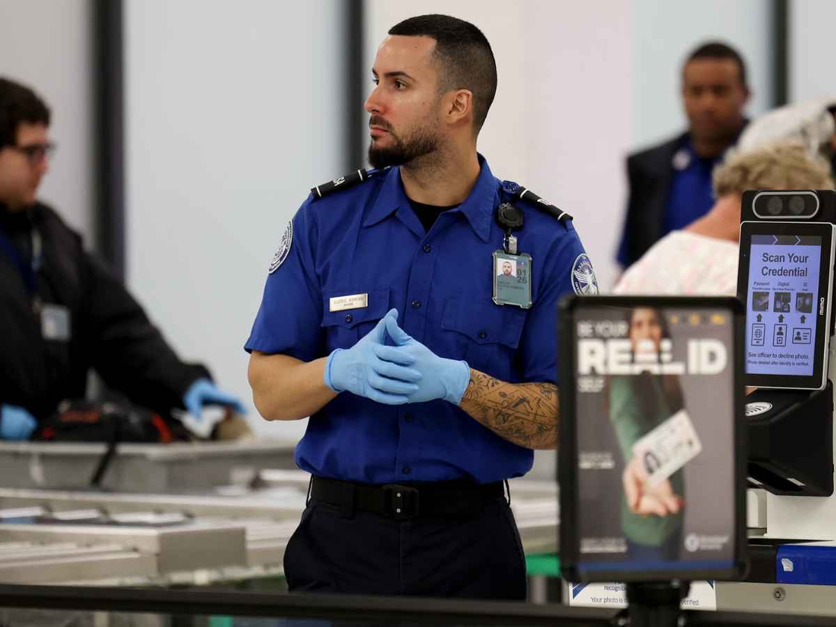 TSA officer at airport security checkpoint with Real ID sign in foreground before 2025 deadline