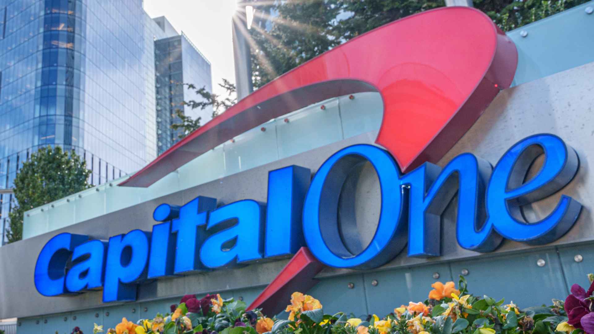 Capital One logo outside its glass headquarters building, surrounded by flowers, ahead of $425 million settlement payouts