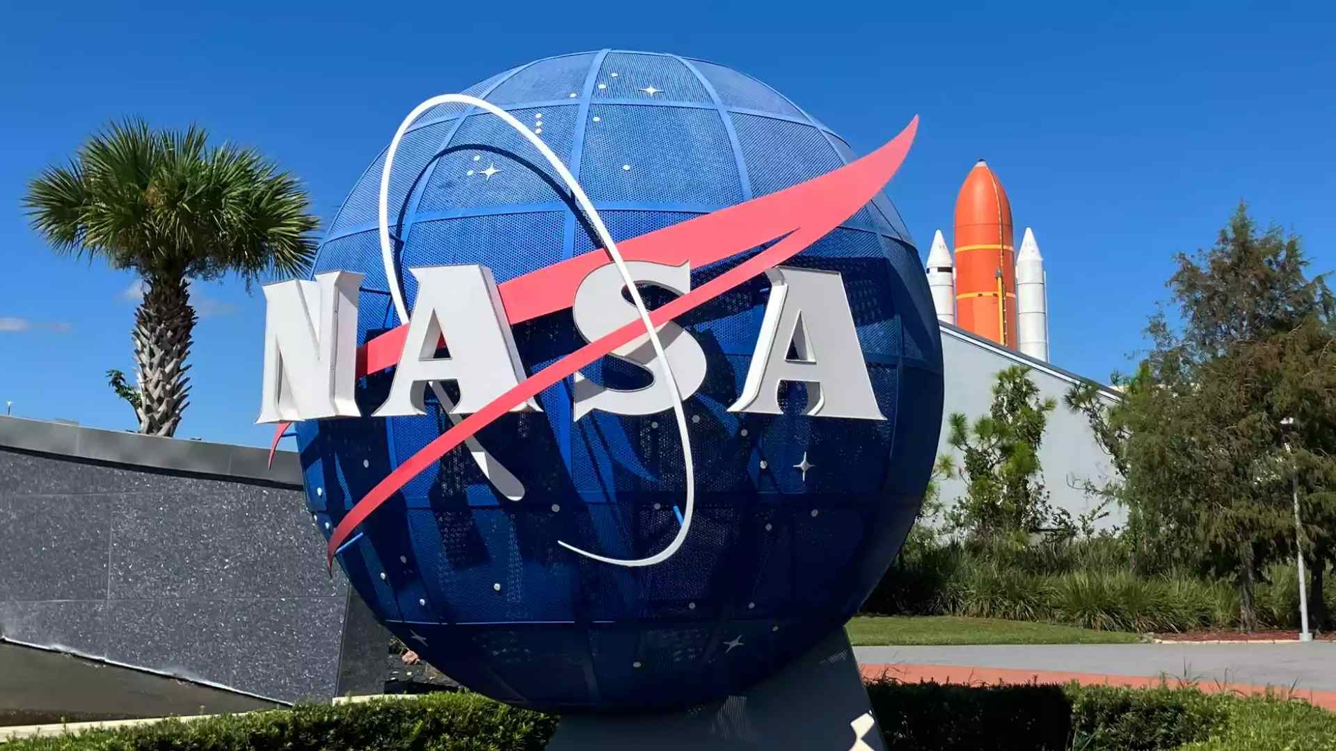 NASA logo in front of a space shuttle display under a clear blue sky at the Kennedy Space Center