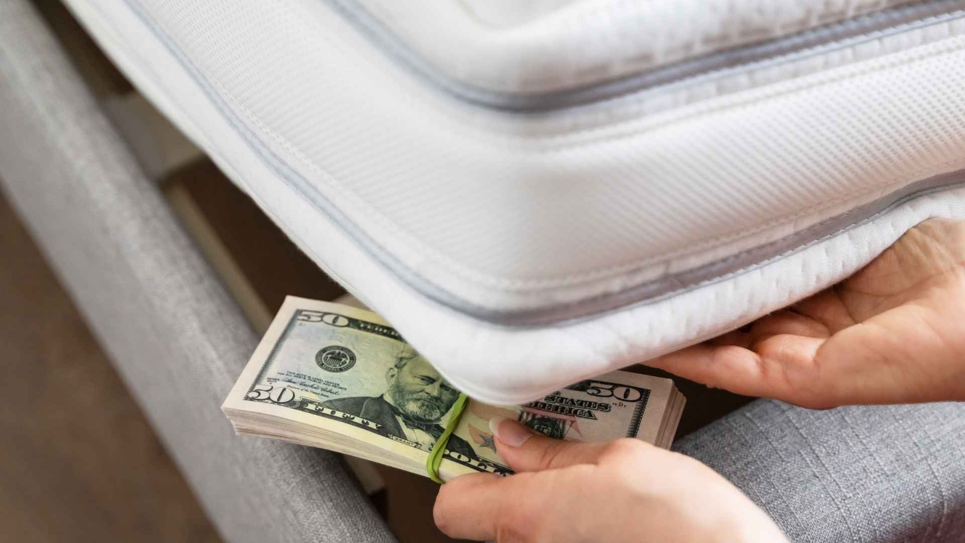 Hands hiding a bundle of fifty-dollar bills under a mattress, illustrating the risks of storing cash at home without protection
