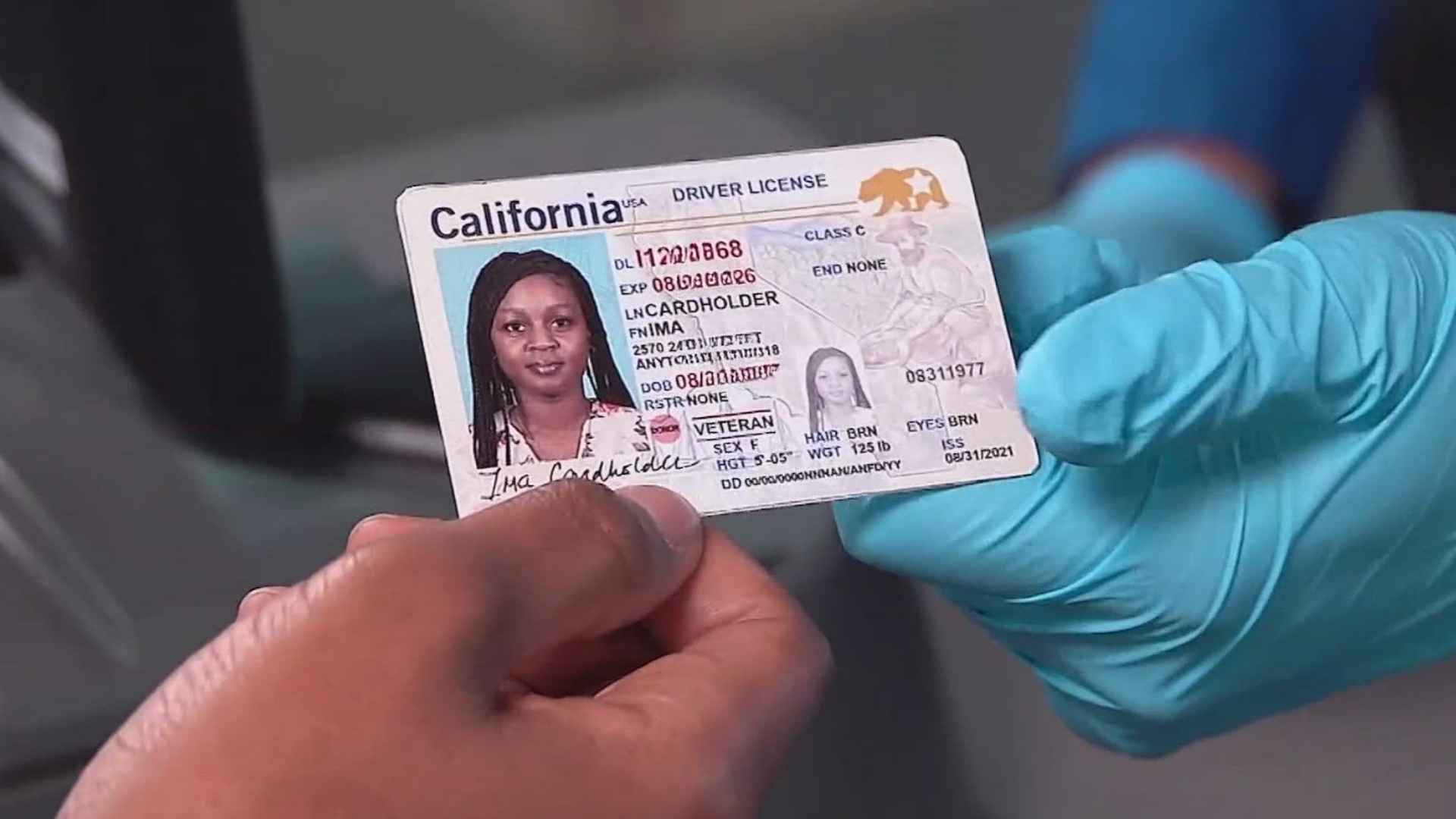 California DMV worker wearing blue gloves hands a Real ID-compliant driver’s license to an eligible resident under the fee-waiver program.