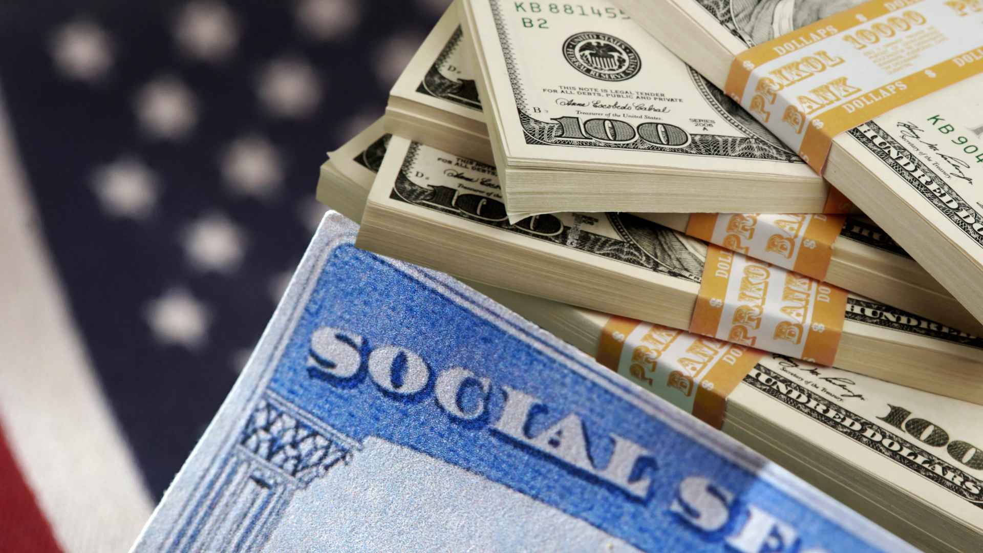 Social Security card beside stacks of hundred-dollar bills against a blurred U.S. flag backdrop.