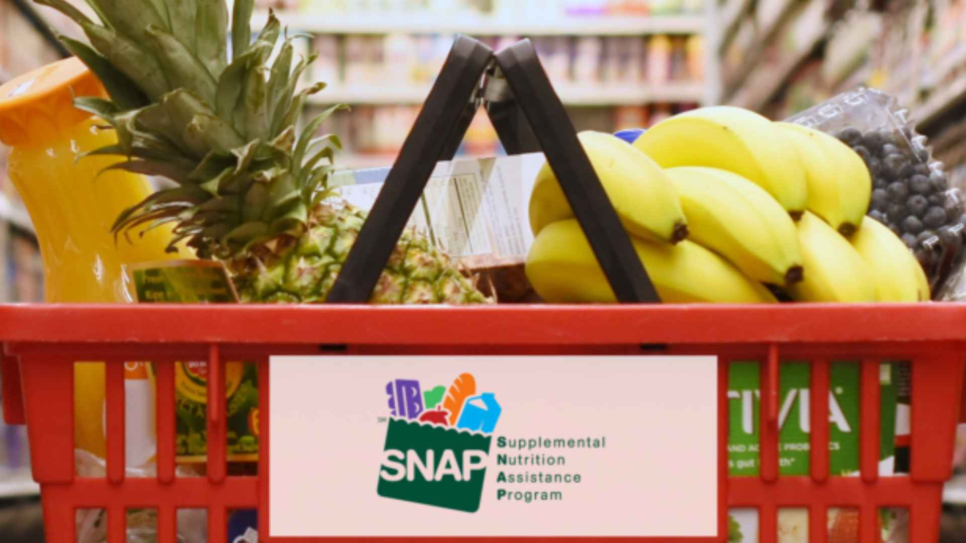Grocery shopping basket with fruits like bananas and pineapple, featuring SNAP (Supplemental Nutrition Assistance Program) logo on the front, in a supermarket aisle