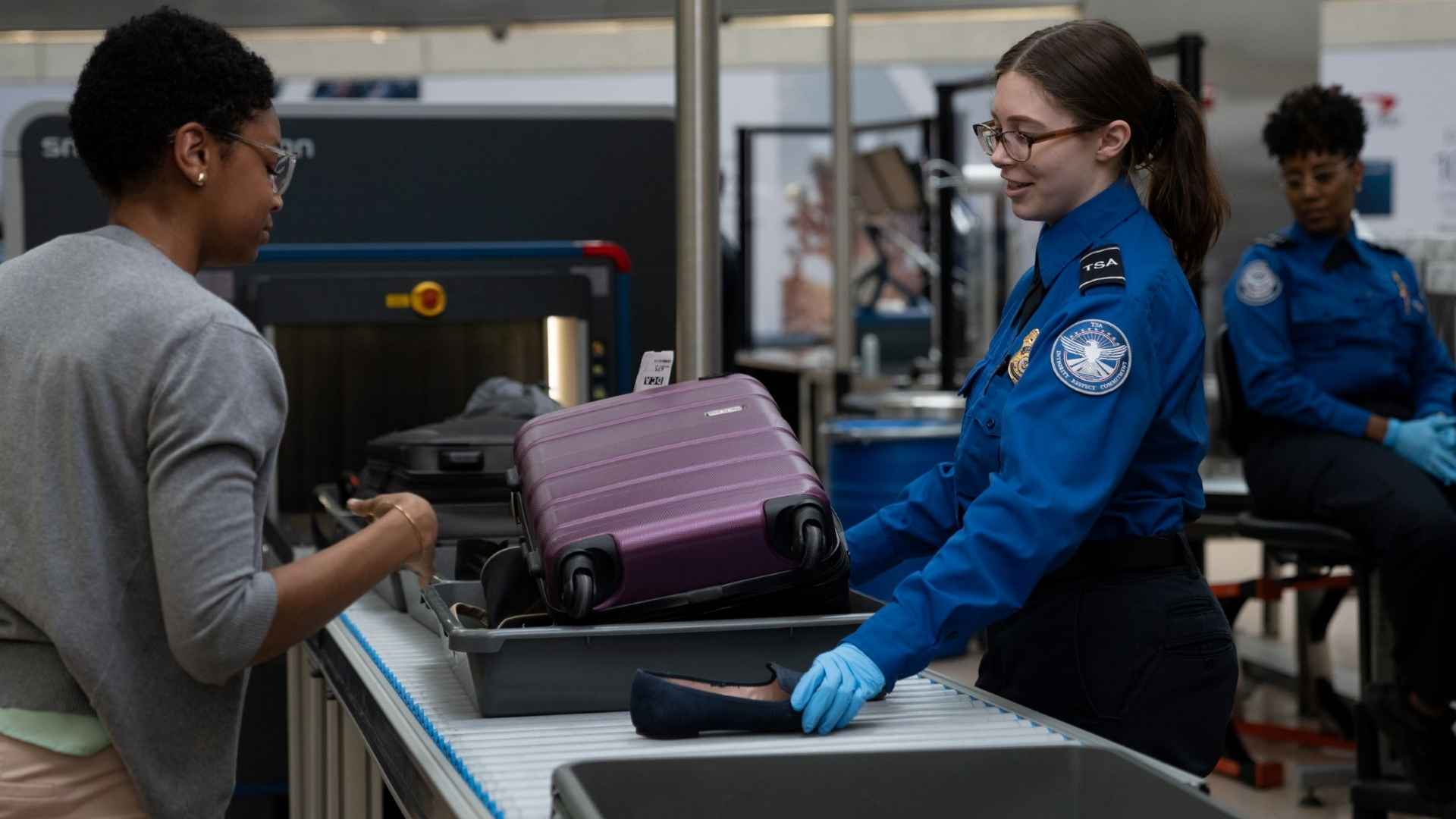 TSA officer inspects carry-on luggage at airport security checkpoint while traveler waits during bag screening process