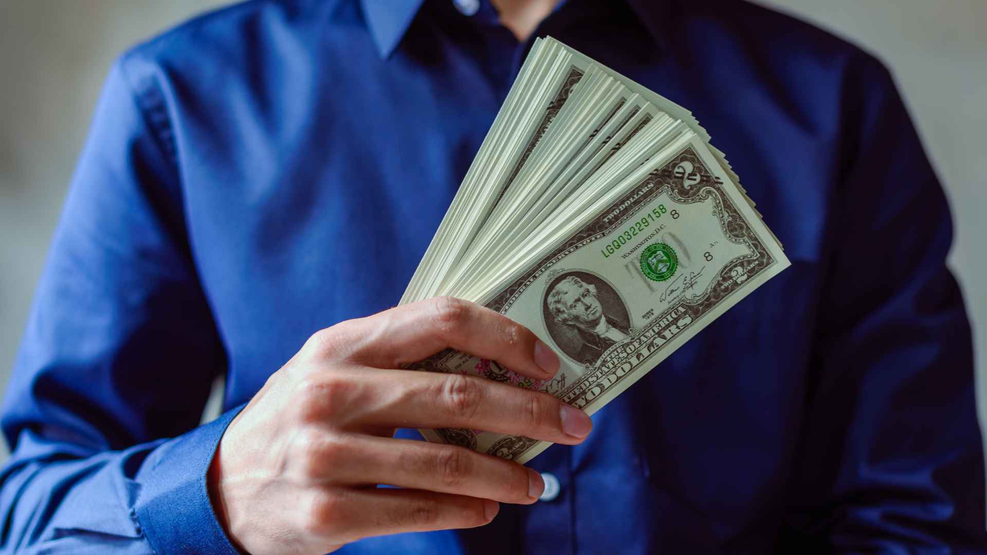 Person in blue shirt holding a fan of two-dollar bills, symbolizing savings gained by filing Social Security form SSA-1020 for Extra Help.