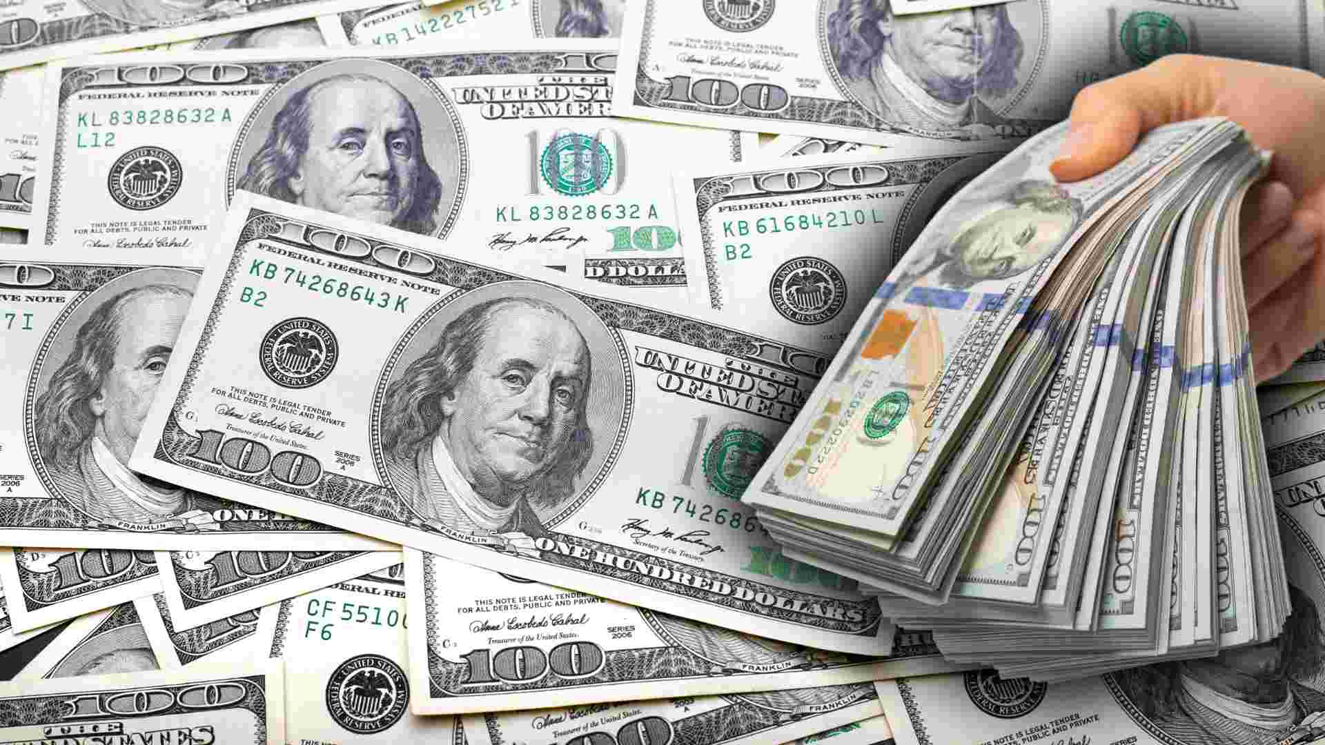 Hand holding a stack of US $100 bills over a background of scattered $100 bills.