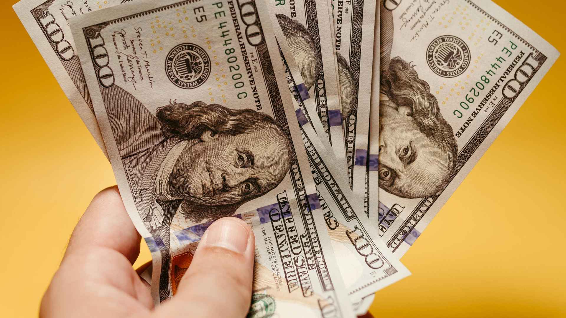 Hand holding several $100 bills on yellow background represents extra cash from combining SSI with ten New York benefits.