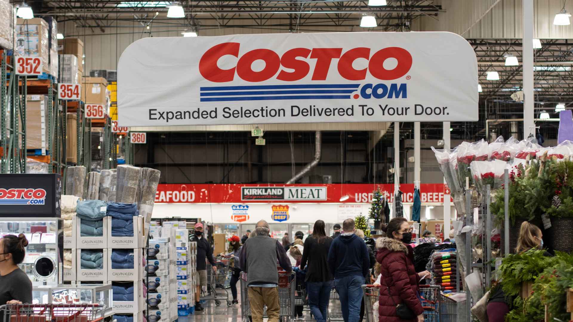 Shoppers inside a Costco warehouse browsing aisles under a banner promoting Costco.com home delivery service