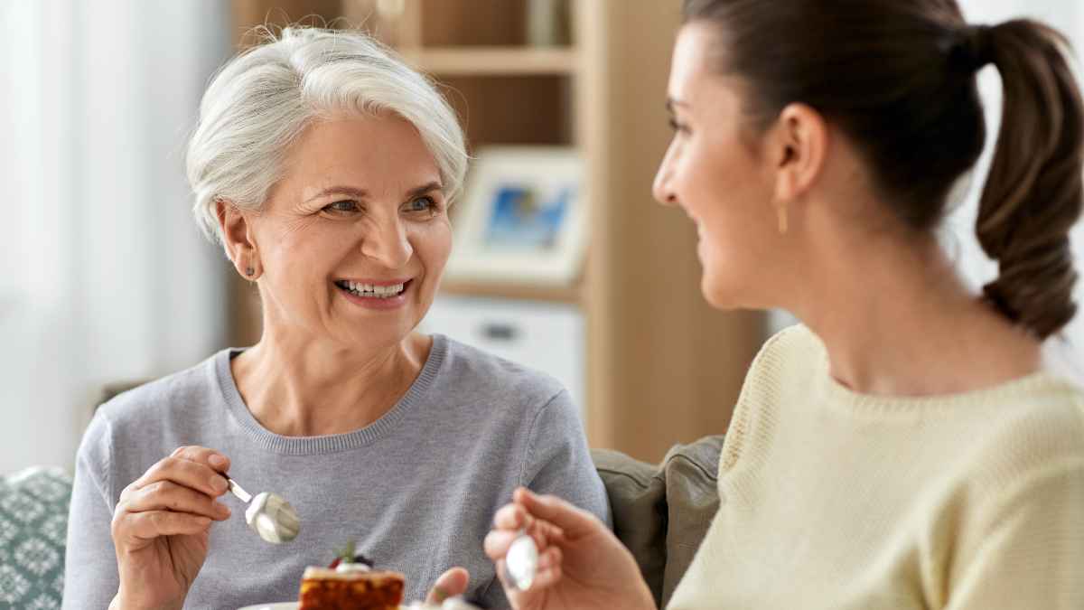 Smiling mother and her adult daughter share dessert on the couch, reflecting parents’ growing financial support for grown children and its impact on retirement plans.