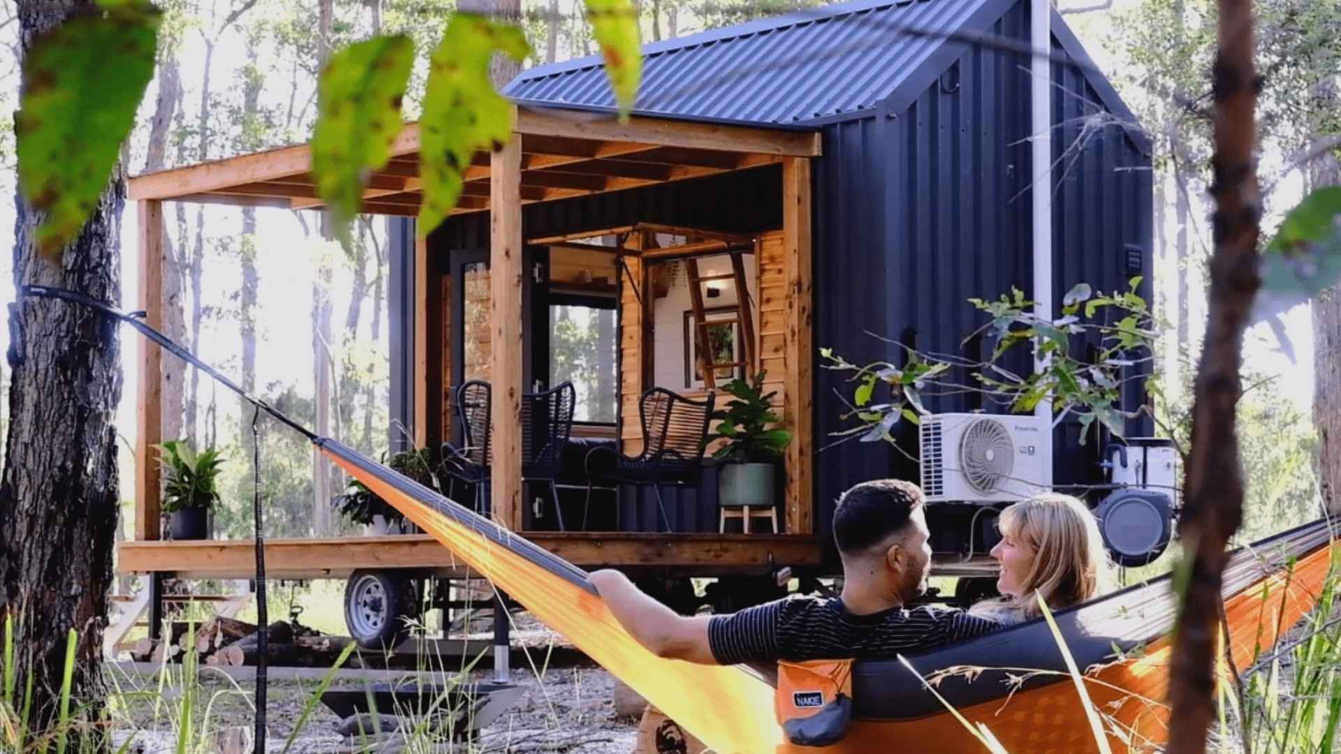 Couple relaxing in a hammock in front of a Gunyah tiny house in a natural setting with trees and greenery.