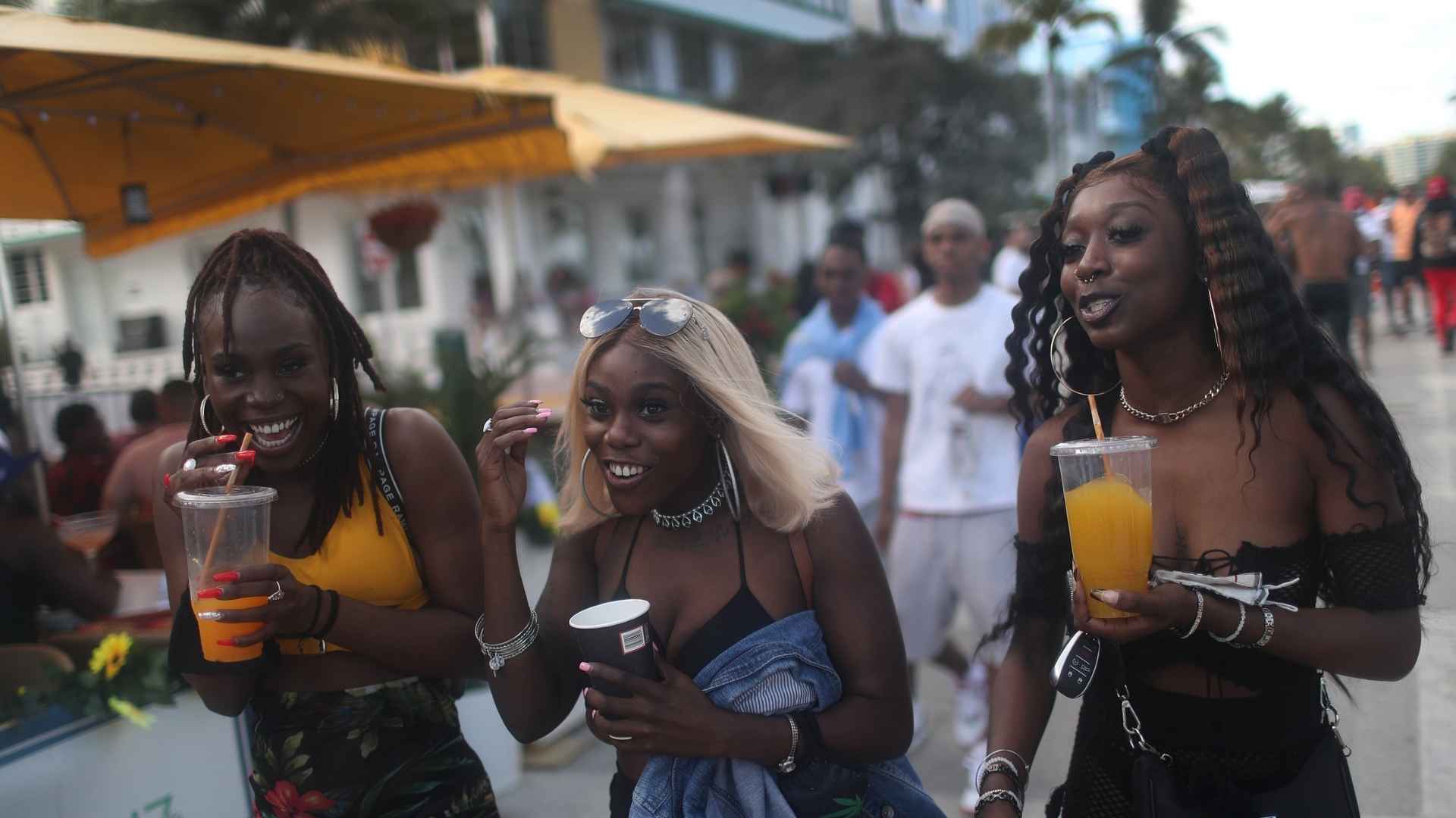 Three women holding beer and cocktail cups while walking on the Miami Beach boardwalk under palm trees.