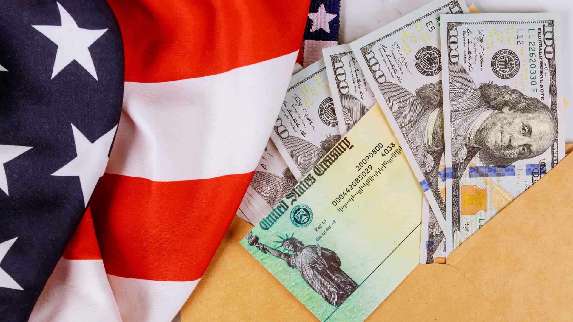 American flag draped next to a U.S. Treasury check and several $100 bills partially tucked into an envelope.