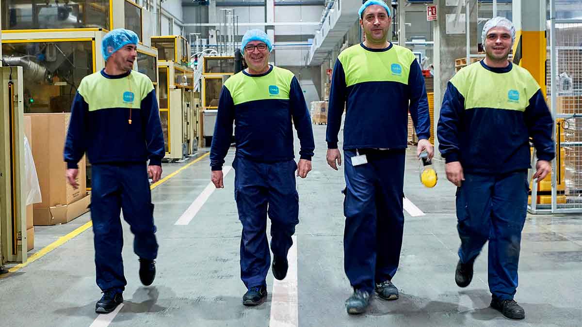 Cuatro operarios de producción con uniforme azul y chaleco amarillo caminan por el interior de la fábrica de Caiba, especializada en envases de plástico, en Las Ventas de Retamosa (Toledo).