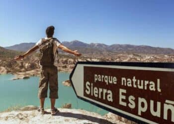 Hombre con mochila contemplando el paisaje en el Parque Natural Sierra Espuña, con un cartel señalando el lugar.