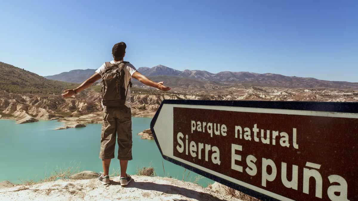 Hombre con mochila contemplando el paisaje en el Parque Natural Sierra Espuña, con un cartel señalando el lugar.