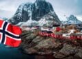 Paisaje de Noruega con casas rojas y bandera noruega ondeando, relacionado con oportunidades de trabajo en piscifactorías de salmón.