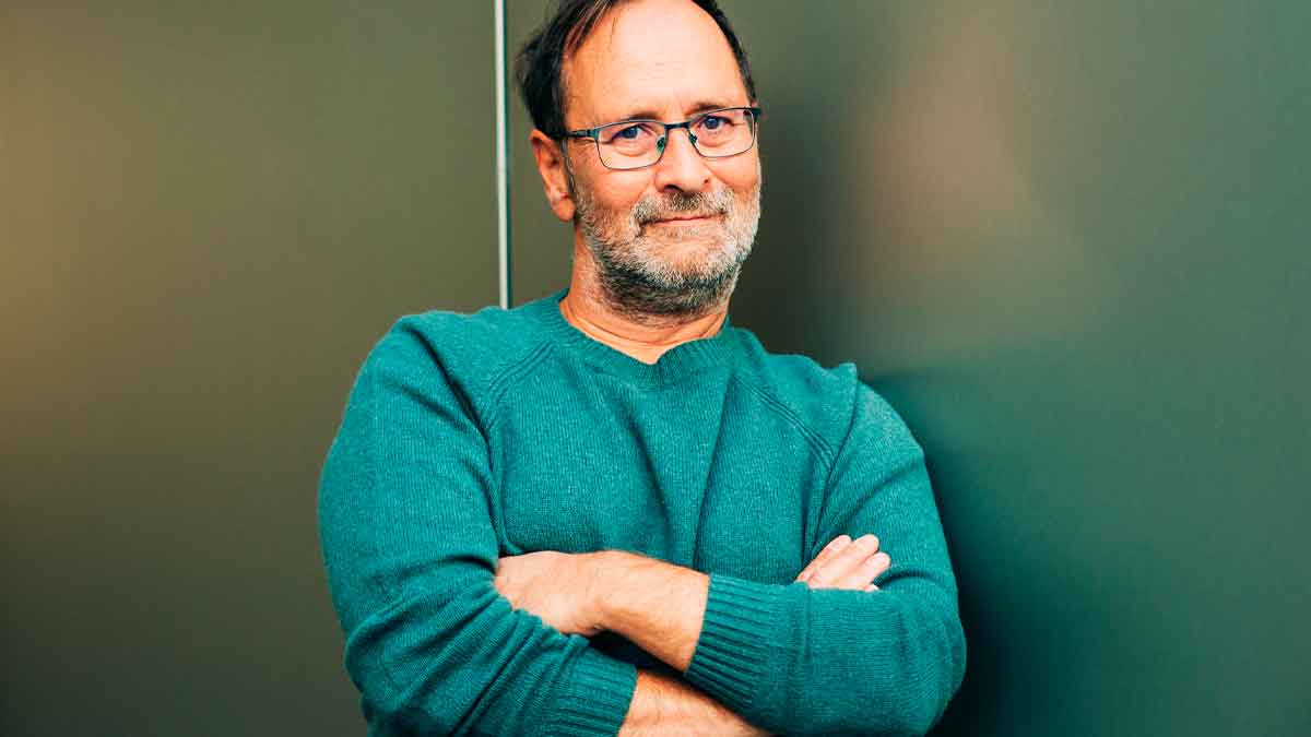 Hombre mayor de 50 años con jersey verde, brazos cruzados y gesto confiado, posible beneficiario de ayudas estatales de 600 € al mes
