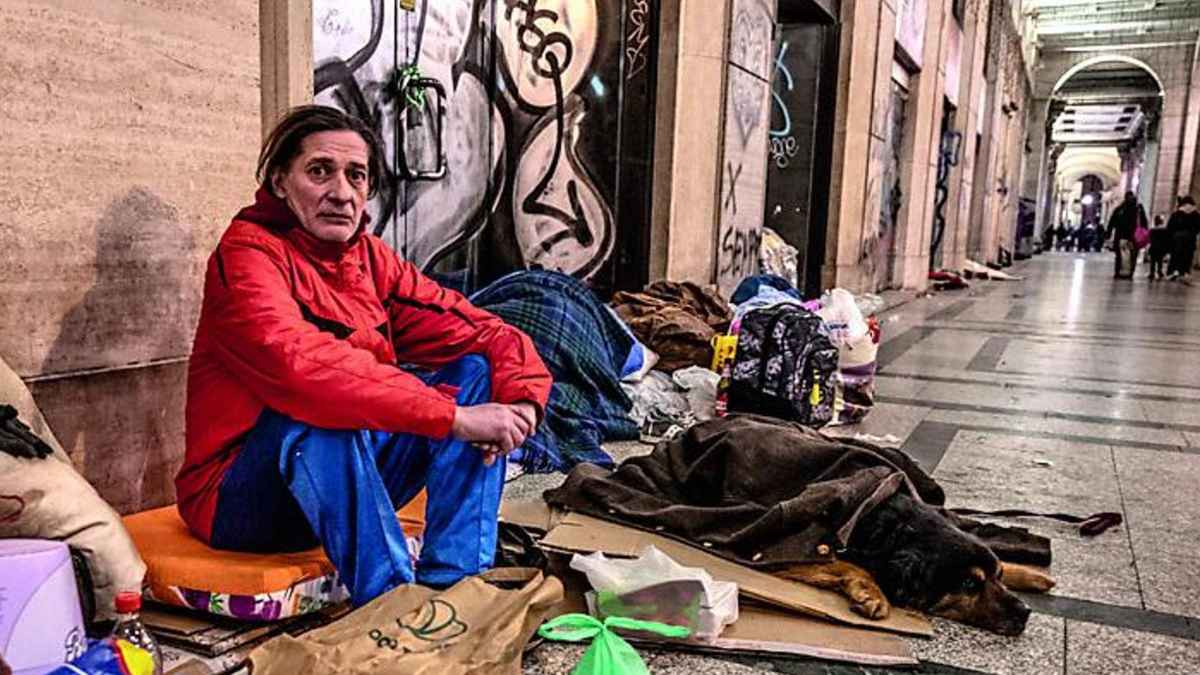 Hombre de 61 años sin hogar sentado en la galería San Federico de Turín junto a su perro tras perder empleo y pensión.