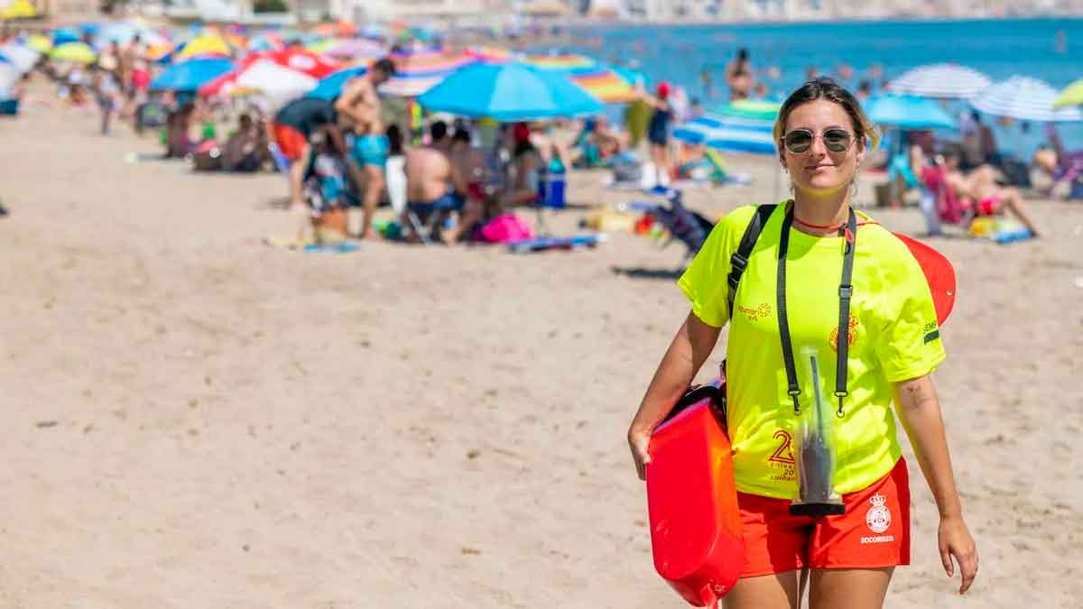 Socorrista profesional con tubo de rescate patrullando una playa llena de bañistas y sombrillas en pleno verano