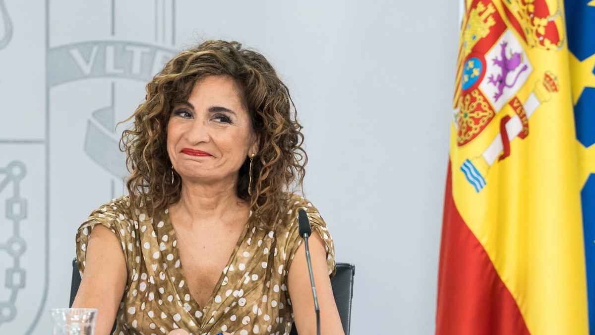 “Ministra de Hacienda María Jesús Montero sonriente en rueda de prensa junto a la bandera de España”