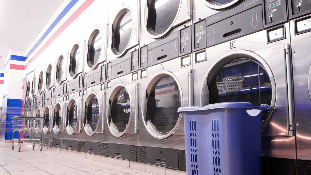 Interior de lavandería autoservicio con cinco lavadoras industriales y cesta de ropa azul