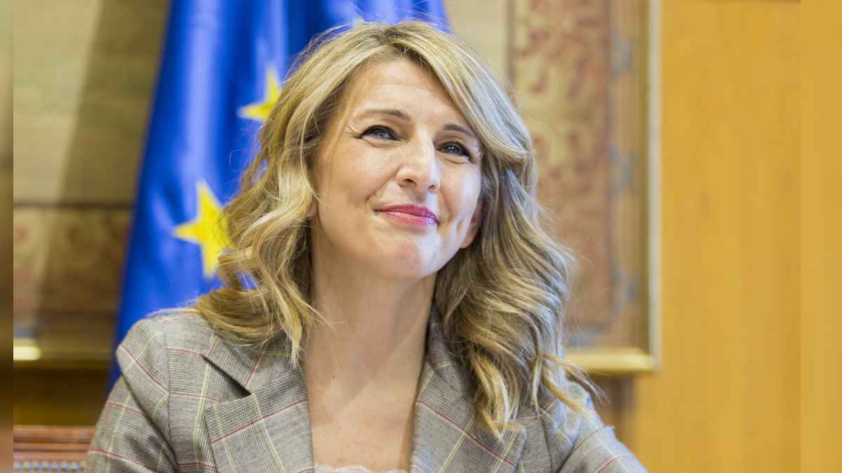 Yolanda Díaz, ministra de Trabajo, sonríe ante bandera de la UE; noticia sobre despido en baja común y descuento del paro por el SEPE