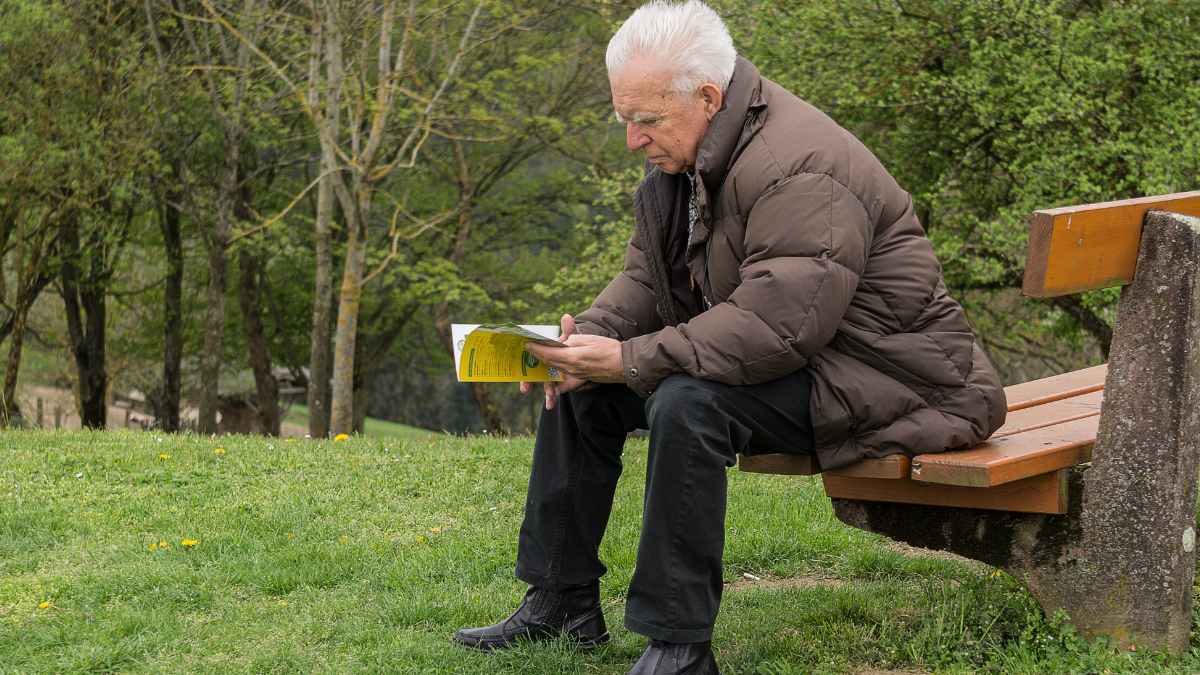 Jubilado sentado en un banco leyendo un folleto, con abrigo de invierno, en un parque