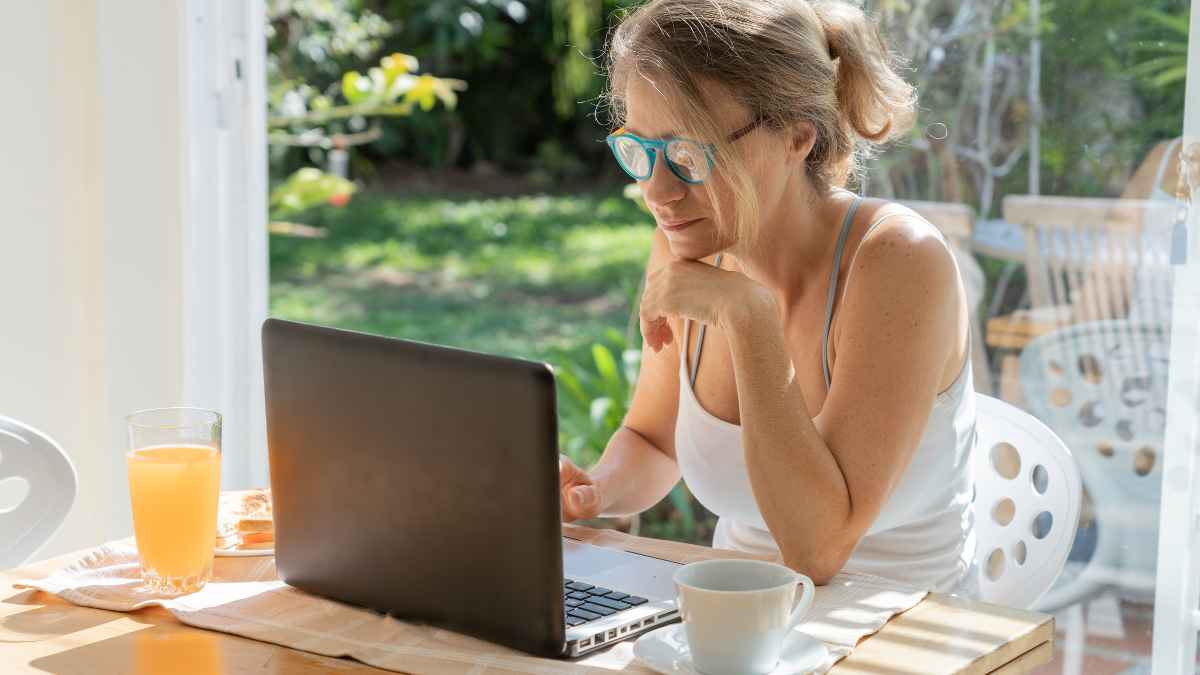 Mujer trabajando desde casa con un portátil frente a la ventana, rodeada de luz natural y un vaso de zumo, ejemplo de cómo el teletrabajo mejora salud y productividad.