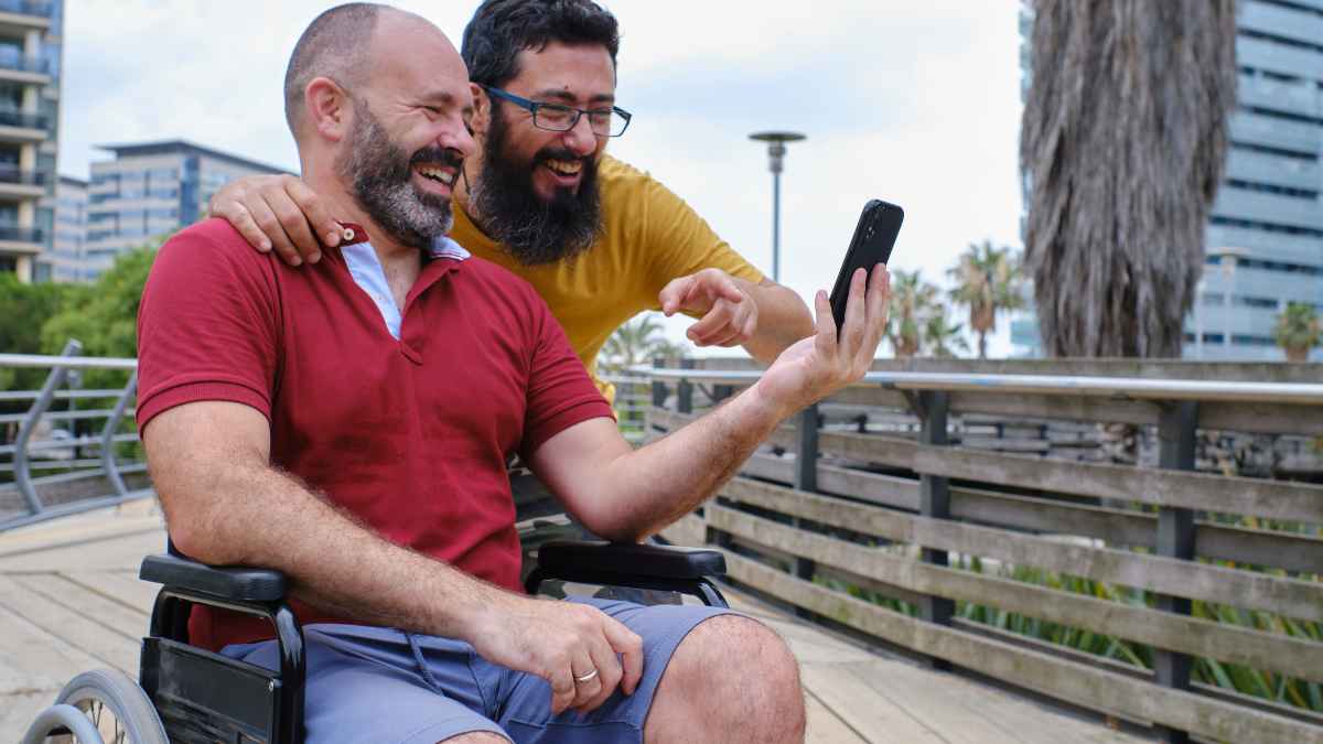 Dos hombres sonríen mirando el móvil, uno de ellos en silla de ruedas, ilustrando las ayudas para personas con discapacidad en Andalucía en 2025.