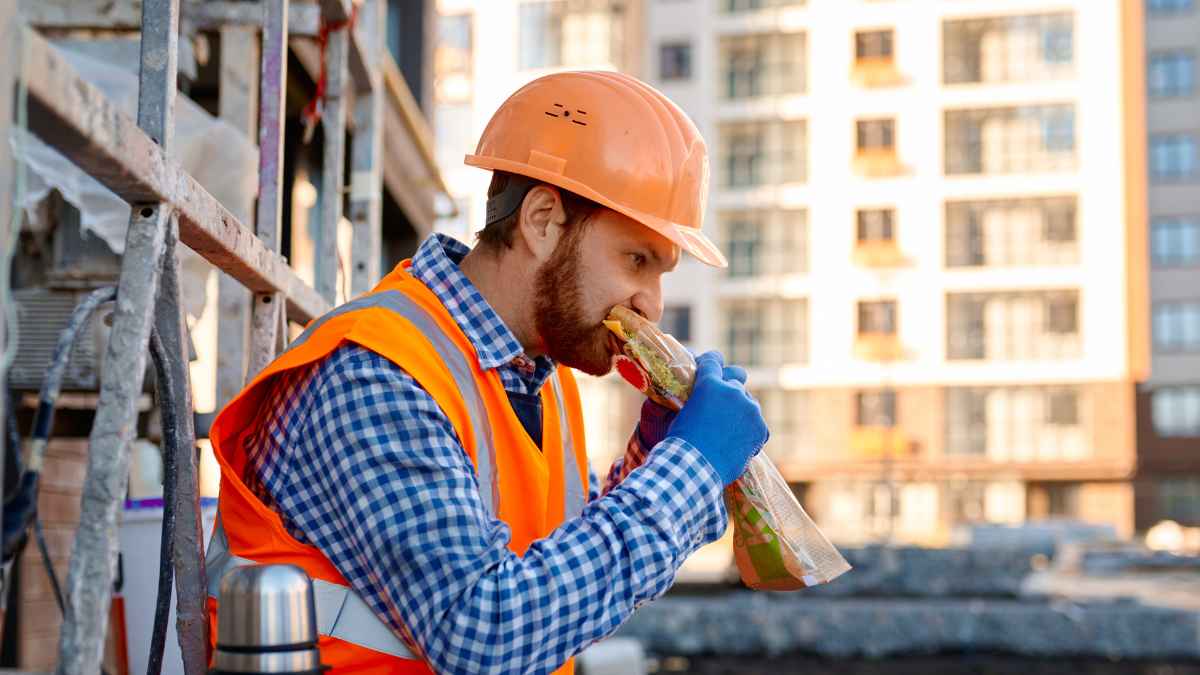 Trabajador de la construcción con chaleco reflectante y casco naranja comiendo bocadillo en su descanso laboral de 20 minutosTrabajador de la construcción con chaleco reflectante y casco naranja comiendo bocadillo en su descanso laboral de 20 minutos