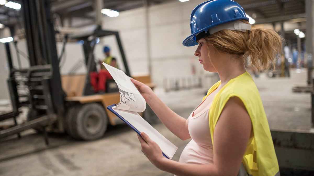 Mujer embarazada trabajando en una construcción con casco y chaleco de seguridad, mientras revisa planos en una obra.