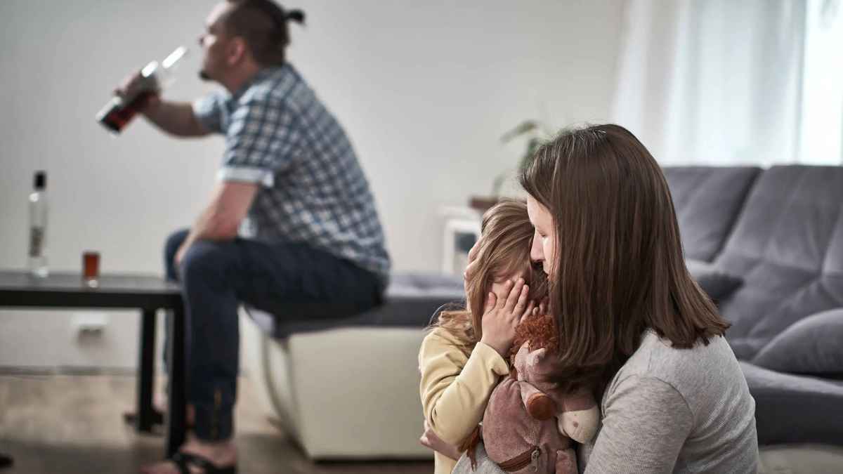 Hombre bebiendo alcohol mientras madre consuela a su hija en casa