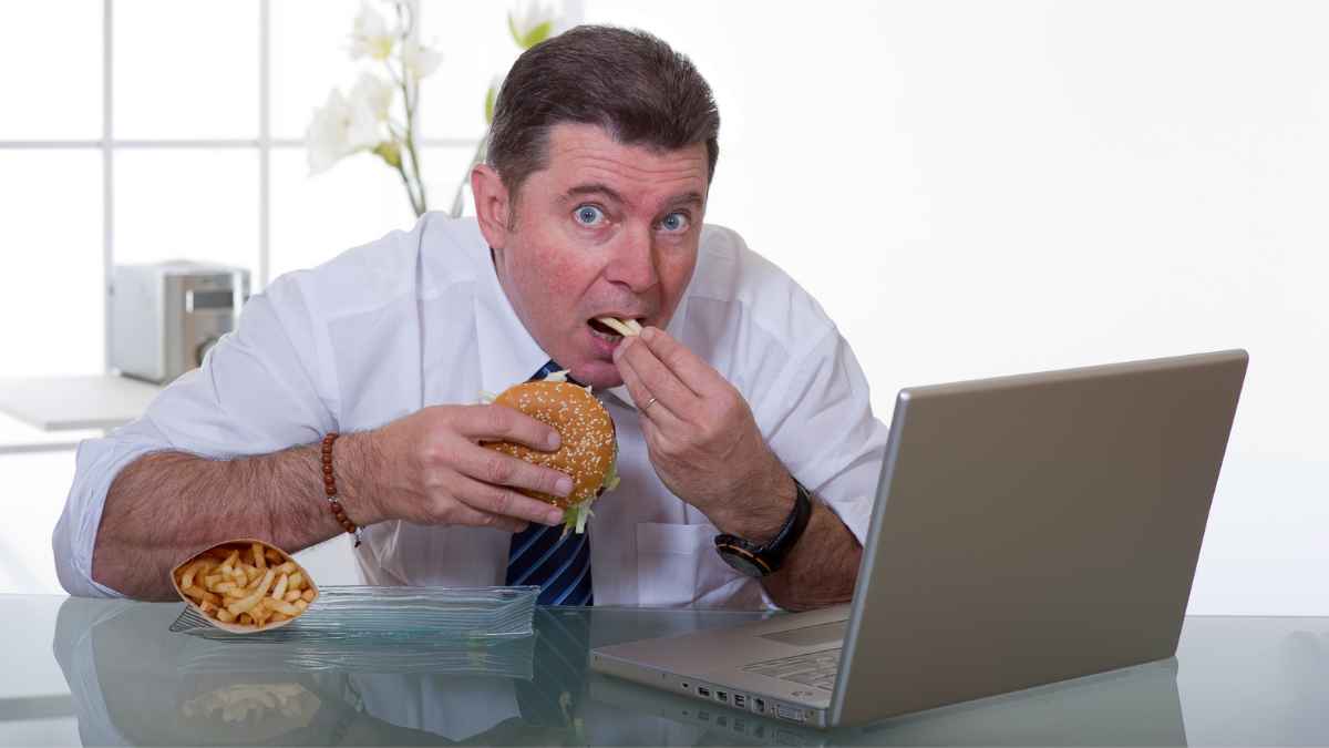 Hombre comiendo hamburguesa y patatas fritas frente al ordenador por estrés laboral