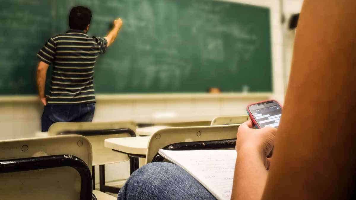 Profesor escribiendo en la pizarra mientras un alumno usa el móvil en clase