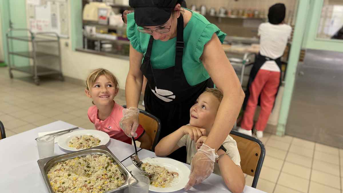 Trabajadora de comedor escolar sirviendo comida a dos niños en un centro educativo.