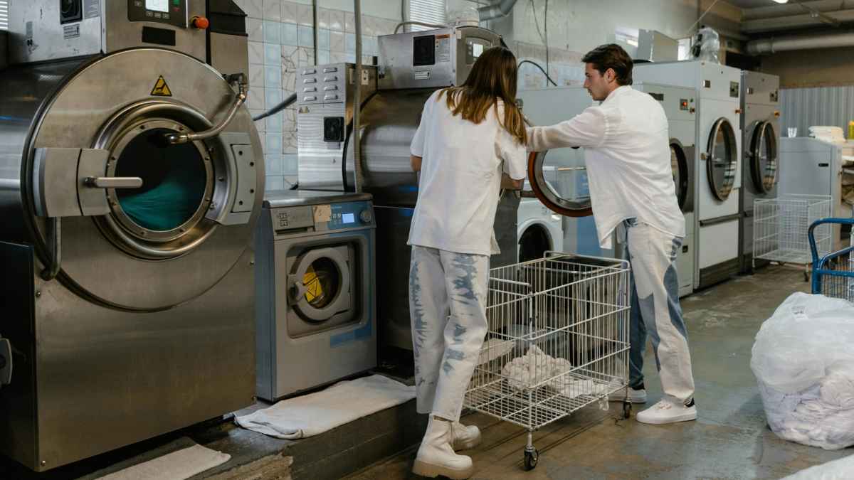 Personas trabajando en lavandería industrial con máquinas de lavado y carros de ropa