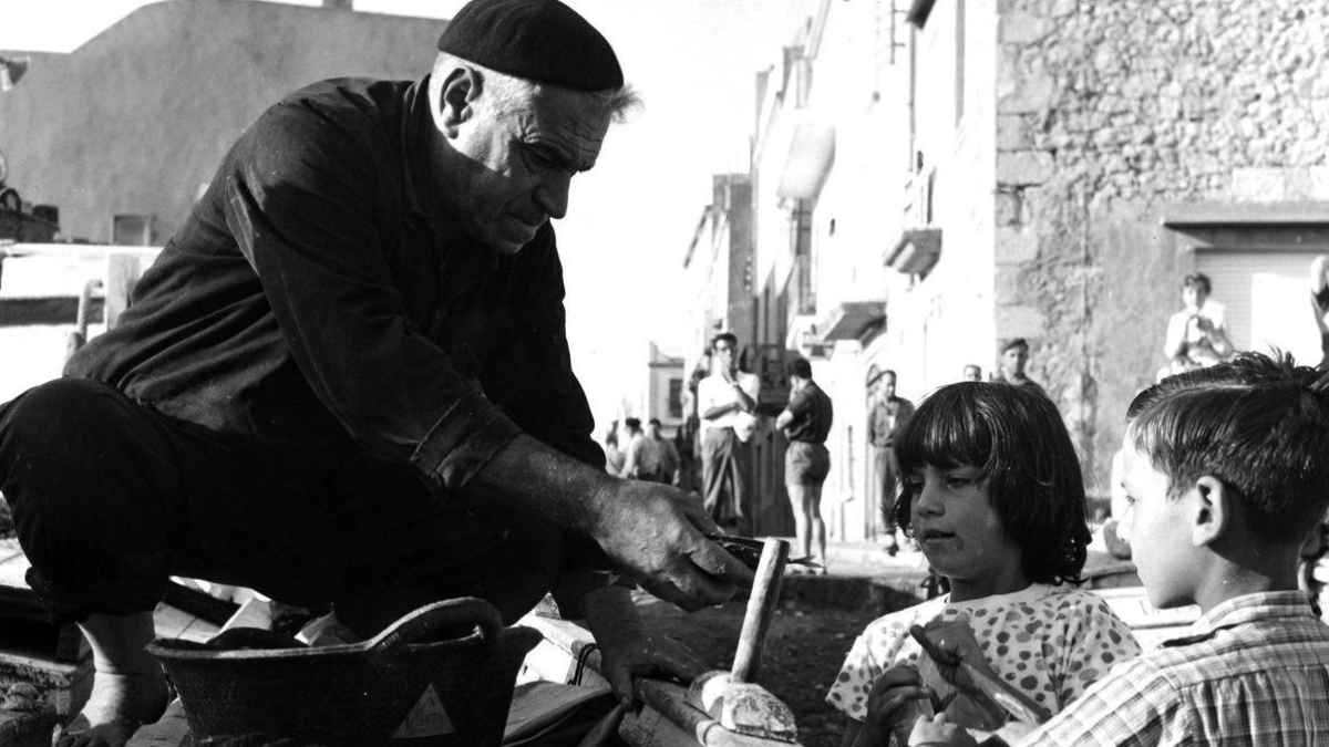 Hombre mayor mostrando utensilio a dos niños en una calle durante la posguerra española